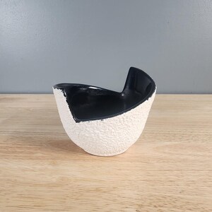 May include: A small, white ceramic planter with a black glazed interior. The planter has a textured, rough exterior and a unique, asymmetrical shape.