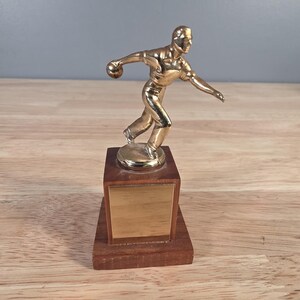 May include: A gold-colored bowling trophy featuring a bowler in mid-throw. The figure is mounted on a wooden base with a light-colored front panel and a darker, rectangular base. The trophy is a classic representation of bowling.