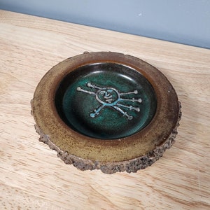 May include: A small, round, brown ceramic ashtray with a green glazed interior. The ashtray has a stylized design of a face with radiating lines.