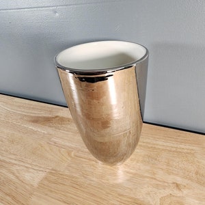 May include: A silver metallic vase with a white interior. The vase is tall and narrow, with a rounded bottom and a slightly flared top.