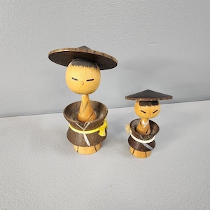 May include: Two wooden Japanese kokeshi dolls with black hats and brown and white painted clothing. The larger doll has a yellow sash.