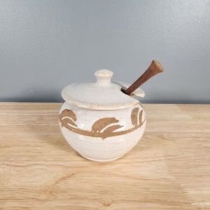 May include: White ceramic honey pot with a brown floral design and a wooden honey dipper. The pot has a lid and is sitting on a wooden surface.
