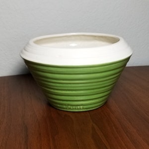 May include: A green and white ceramic planter with a ribbed design. The planter is empty and sitting on a wooden surface.