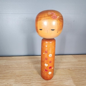 May include: A wooden Japanese kokeshi doll with a painted face and floral pattern. The doll has a round head and a long, slender body.