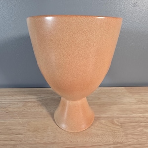 May include: A peach-coloured ceramic vase with a speckled texture. The vase has a wide, flared top and a pedestal base. The vase is on a wooden surface against a grey background. The vase is a decorative item.
