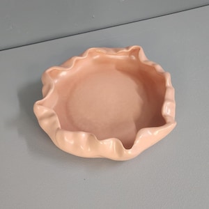 May include: A pale pink ceramic bowl with a ruffled edge. The bowl is round and shallow, with a smooth, glossy finish.