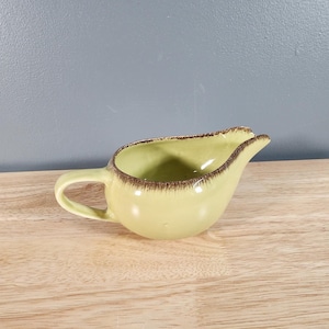 May include: A small, green ceramic creamer with a brown rim. The creamer has a rounded shape and a small handle.