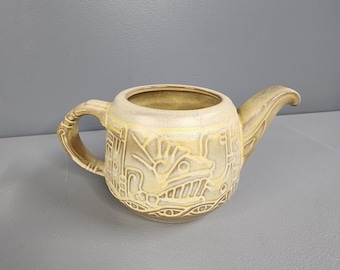 May include: A ceramic teapot with a brown and yellow glaze. The teapot has a stylized design of a bird or animal on the side. The teapot has a handle and a spout.