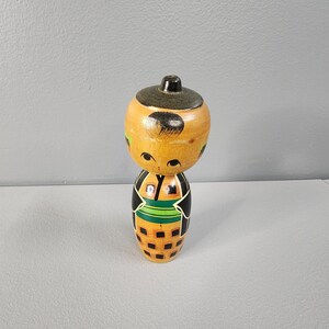 May include: A wooden Japanese Kokeshi doll with a round head and cylindrical body. The doll features a light brown head with black hair and facial details. The body is decorated with black, green, and orange geometric patterns. The doll is approximately 6 inches tall.