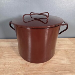 May include: A deep, cylindrical stockpot with a glossy, dark brown enamel finish. The pot features two curved side handles and a matching lid with a distinctive, elevated cross-shaped metal handle centered on top for easy lifting and secure closure.