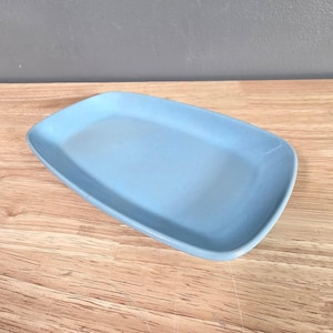 May include: A light blue rectangular ceramic serving tray with rounded corners. The tray is empty and sits on a wooden surface. The background is a solid gray color.