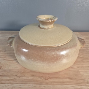 May include: A ceramic lidded casserole dish with a round shape and a light brown and tan colour gradient. The lid has a small handle, and the dish has two side handles. The dish is on a wooden surface.