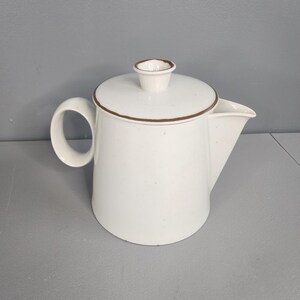 May include: White ceramic coffee pot with a brown rim and handle. The pot has a lid with a small knob and a spout for pouring. The coffee pot is a classic design, perfect for serving coffee or tea.