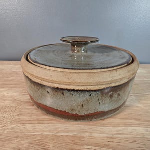 May include: A handmade ceramic lidded bowl with a neutral color palette. The bowl features a round shape with a lid and a small handle. The exterior has a mix of beige, gray, and brown tones, with a band of red near the base.