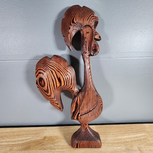 Pode incluir: Escultura de galo em madeira com um design em camadas. A escultura é principalmente castanha com detalhes em laranja, apresentando uma crista, bico e cauda detalhados. O galo está sobre uma pequena base quadrada, mostrando um estilo artístico único.