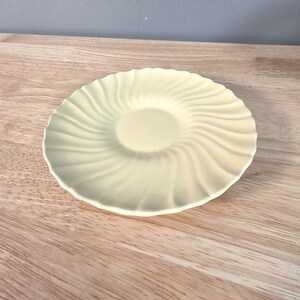May include: A pale yellow saucer with a spiral design. The saucer is round with a slightly raised center and a rippled edge. The saucer is made of ceramic or porcelain.