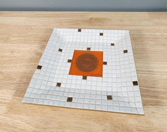 Puede incluir: Una bandeja de cerámica blanca con un inserto cuadrado naranja que presenta un diseño geométrico marrón. La bandeja está hecha de pequeñas baldosas blancas con acentos negros.
