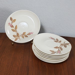 May include: A set of eight white ceramic saucers with a brown and pink floral design. The saucers are stacked in a pile on a wooden surface.