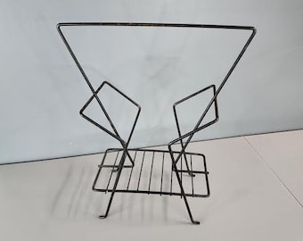 May include: A black metal magazine rack with a geometric design. The rack has a rectangular base and two triangular sides that meet at the top. The rack is made of thin metal rods that are welded together.