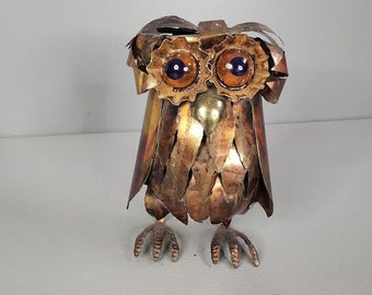 May include: A copper owl sculpture with blue eyes and a textured surface. The owl is made of metal and has a rustic look.