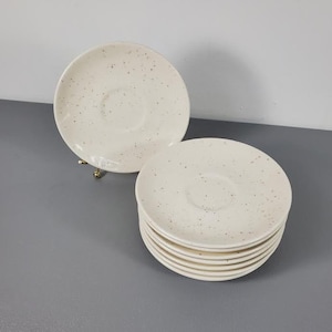 May include: A stack of eight white ceramic saucers with a speckled pattern. The saucers are perfect for serving coffee or tea.