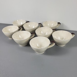 May include: A set of eight cream-colored teacups with speckled detailing. Each cup has a dark brown handle. The cups are arranged in a slightly offset circular pattern, showcasing their design and form. Ideal for serving tea or coffee.