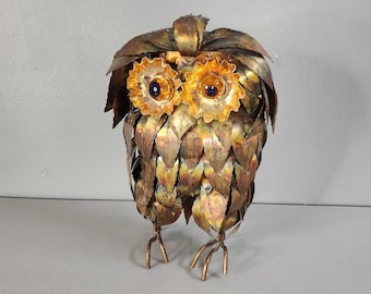 May include: A copper owl sculpture with a textured, scaly body and large, round eyes with blue pupils. The owl is standing on two legs with three toes each.