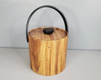 May include: A wooden ice bucket with a black leather handle and a black lid. The bucket has a wood grain pattern.