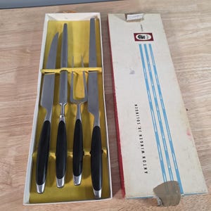 May include: A vintage stainless steel carving set with black handles, including a carving knife, a slicer, and a carving fork, presented in a yellow-lined box. The box is open, revealing the set, and the lid is partially visible.