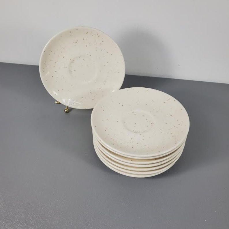 May include: A stack of eight white ceramic saucers with a speckled pattern. The saucers are perfect for serving coffee or tea.