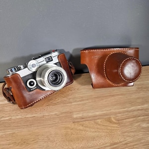 May include: A vintage Argus C3 camera with a brown leather case and strap. The camera has a silver lens and body with black accents. The leather case is open, revealing the camera. The camera is on a wooden surface.