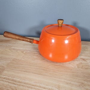 May include: An orange enameled saucepan with a wooden handle and lid. The handle and lid knob are made of wood. The saucepan is sitting on a light-colored wooden surface.