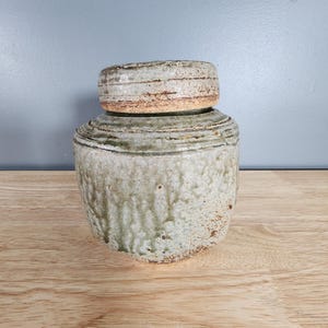 May include: A ceramic jar with a lid, featuring a light green glaze with a textured surface. The jar has a rounded shape and a wide mouth.