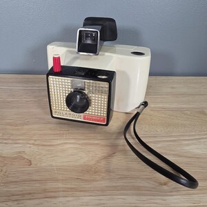 May include: A vintage Polaroid Swinger Land Camera, model 20, with a black and gold lens and a red button. The camera is black and gold with a white body and a black viewfinder. A black wrist strap is attached.