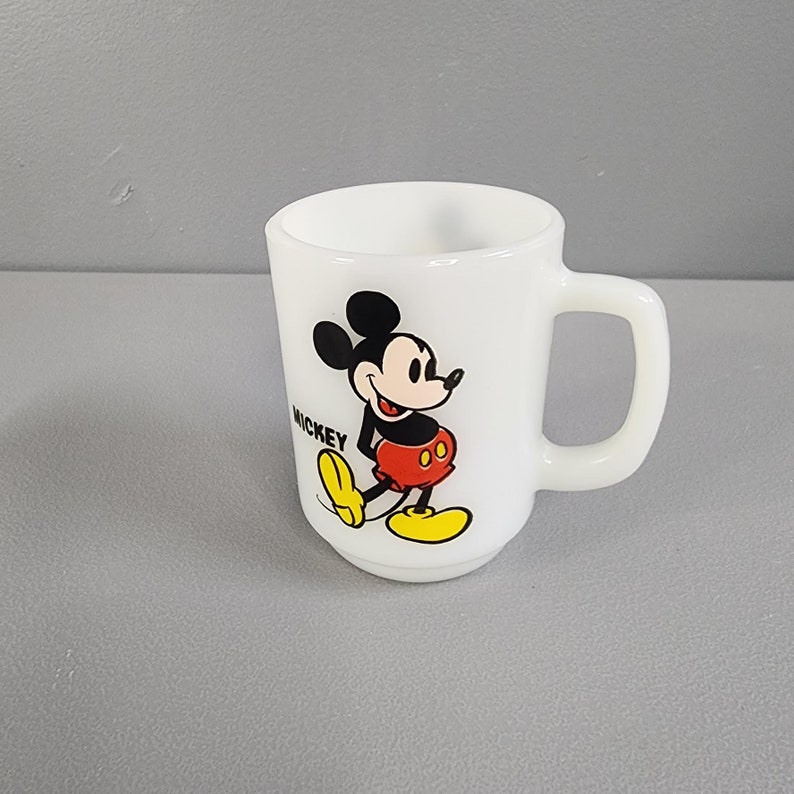 May include: White ceramic mug with a cartoon image of Mickey Mouse in red shorts and yellow shoes. The text "MICKEY" is printed on the mug.