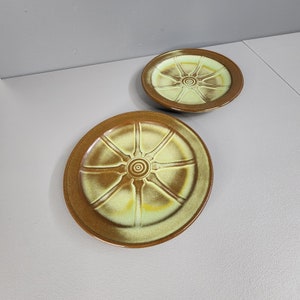 May include: Two green and brown ceramic plates with a wheel design. The plates are slightly different shades of green and brown.
