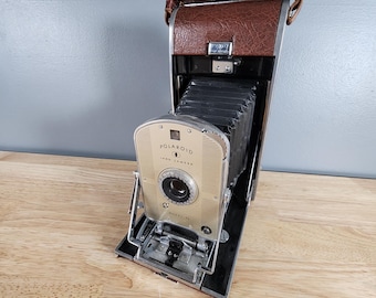 Puede incluir: Una cámara Polaroid Land vintage con una funda de cuero marrón y un cuerpo de metal plateado. La cámara tiene una lente negra y un flash de metal plateado. La cámara está en buen estado y tiene un aspecto clásico.