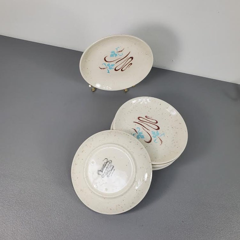 May include: Set of six white ceramic dessert plates with a speckled finish. The plates feature a blue and brown floral design with the text "Lenox" in a brown rectangle.
