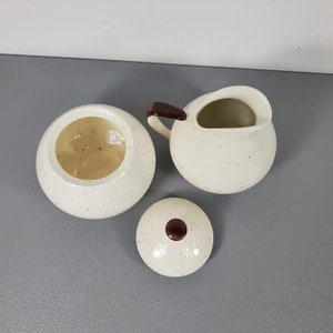May include: A white ceramic sugar bowl and creamer set with brown accents and a speckled finish. The creamer has a handle and a spout. The sugar bowl has a lid with a brown knob.