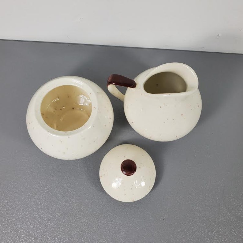 May include: A white ceramic sugar bowl and creamer set with brown accents and a speckled finish. The creamer has a handle and a spout. The sugar bowl has a lid with a brown knob.