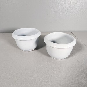 May include: Two white ceramic plant pots with a slightly raised rim. The pots are empty and sitting on a light gray surface.