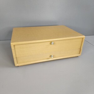 May include: A light wood two-drawer box with silver knobs. The box is closed and sitting on a gray surface.