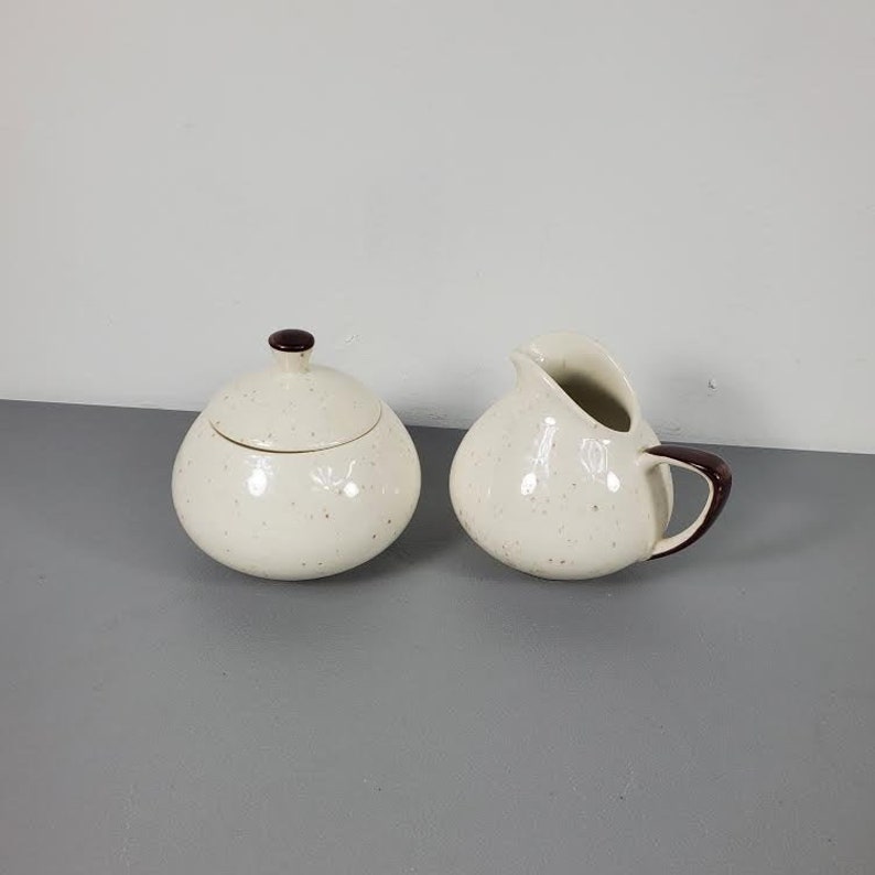 May include: A white ceramic sugar bowl and creamer set with brown trim and a speckled finish.