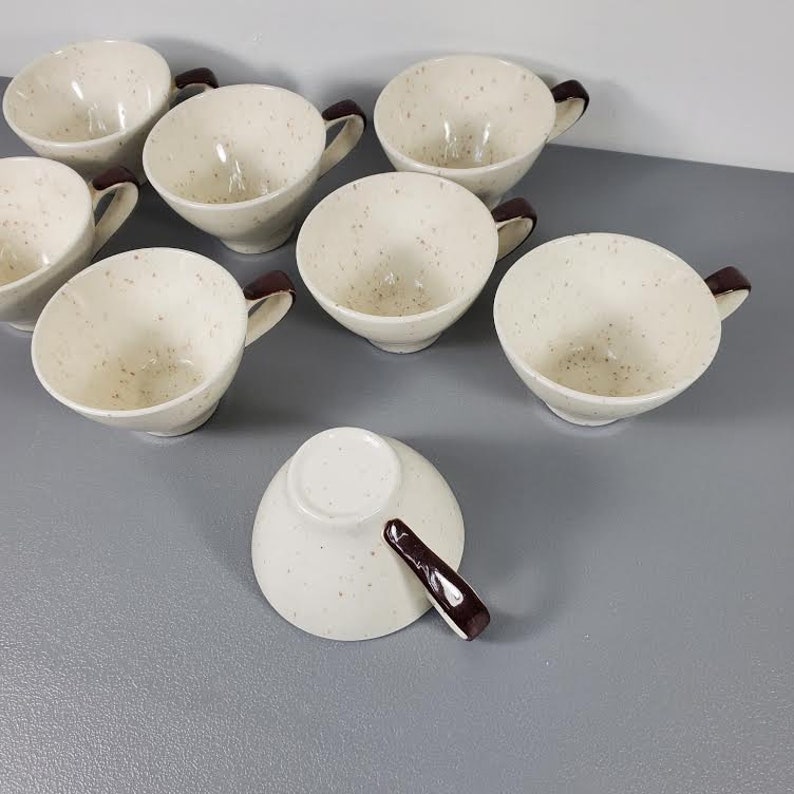Puede incluir: Un conjunto de ocho tazas de cer&aacute;mica color crema con detalles moteados. Cada taza tiene un asa marr&oacute;n oscuro. Las tazas est&aacute;n dispuestas sobre una superficie gris, con una taza boca abajo.