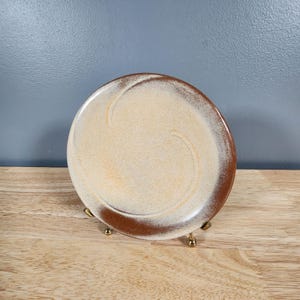 May include: A small, round, ceramic plate with a brown rim and a white center. The plate has a swirl design in the center.