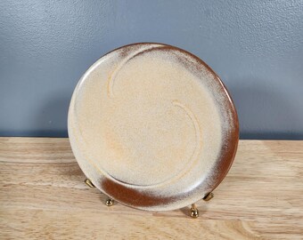 May include: A small, round, ceramic plate with a brown rim and a white center. The plate has a swirl design in the center.