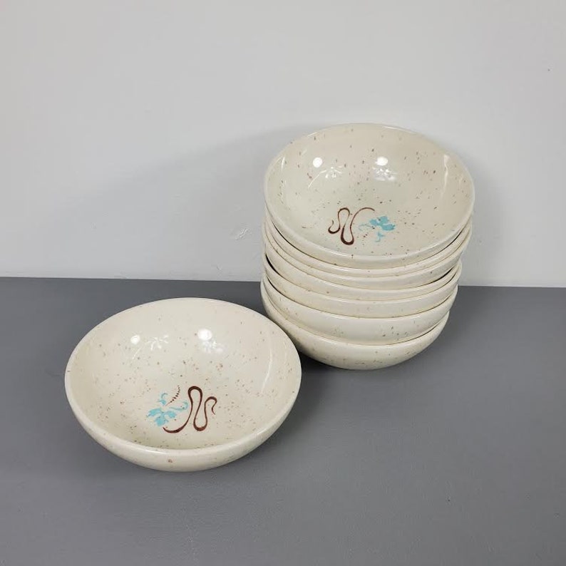 May include: A set of eight white ceramic bowls with a speckled brown and white glaze. Each bowl features a blue and brown floral design.