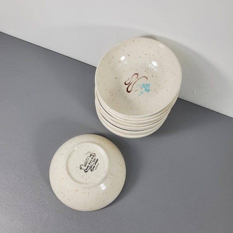 May include: A stack of 8 white ceramic bowls with a speckled pattern. The top bowl has a blue and brown floral design. The bottom bowl has a white label with the text "Fiestaware, Fine China, Made in USA".