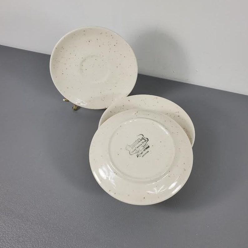 May include: Three white ceramic saucers with a speckled pattern. The saucers have a small indentation in the center. The bottom of one saucer has the text "Made in England, Roseville Pottery, Staffordshire" printed on it.