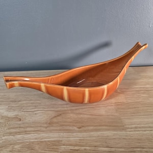 May include: A mid-century ceramic serving dish in a warm orange hue, featuring vertical cream stripes. The dish has a long, boat-like form with a handle at each end. It is displayed on a wooden surface.